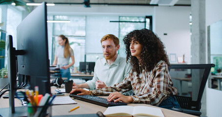 Caucasian female with curly hair typing on keyboard. Colleague with red hair talking to woman. Discussing plans for improvement and possible financial situation.