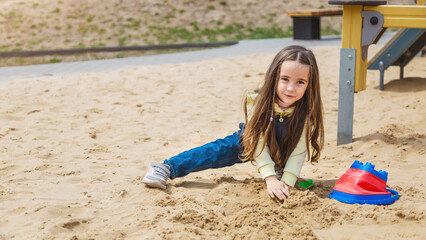 Pretty child girl playing in sand on outdoor playground. Beautiful baby in jeans trousers having fun on sunny warm summer day. Child with colorful sand toys. Healthy active baby outdoors plays games