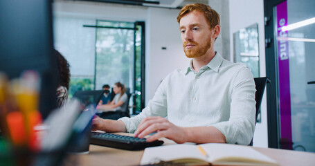 Fototapeta premium Caucasian man red hair working in busy office. Typing on keyboard. Writing code or message to someone. Noting something down on small notepad for later. Busy hour. Workplace.
