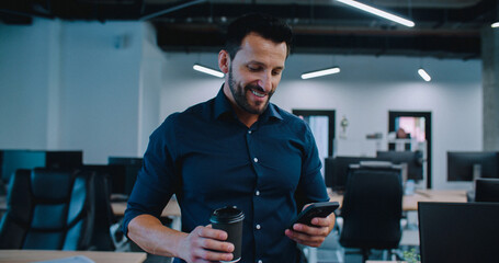 Handsome Caucasian male with dark hair and beard walking down big office space. Holding cup with hot drink with one hand and checking notifications on his phone and smiling.