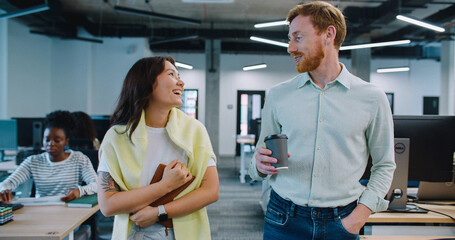 Obraz premium Cute couple walking through their workspace. Casually talking and smiling while enjoying conversation. Discussing possible plans for evening during coffee break. Modern room. Many desks with monitors.