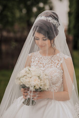 A bride is wearing a veil and holding a bouquet of white flowers. She is smiling and looking at the...