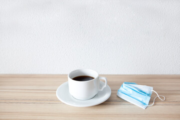 White cup of black coffee with  protective medical mask on wooden  table with empty white background on top