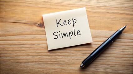 A white sticky note with "keep it simple" written in black marker against a clean wooden desk, surrounded by empty space and minimal distractions.