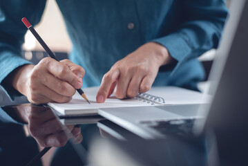 Business woman writing ideas on notebook, using laptop computer searching the information, planning, working at modern office. 
