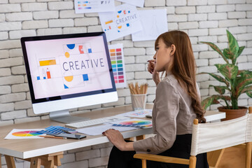 Asian female woman graphic designer focused on creative project in modern office with desktop computer displaying colorful designs