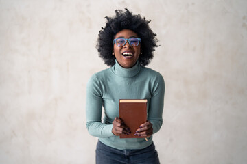 An enthusiastic black woman in stylish glasses and casual attire holding a book, standing before a...