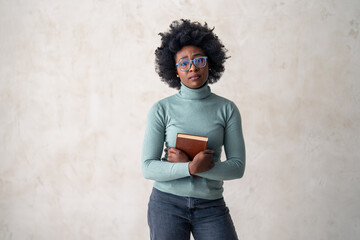 A poised black female with glasses holds a book, wearing a turquoise turtleneck, standing against a...