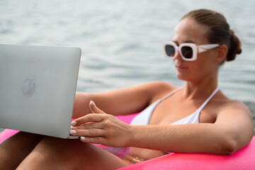 A woman is sitting in a pink inflatable raft on a lake, holding a laptop. Concept of relaxation and leisure, as the woman enjoys her time in the water while working on her laptop.