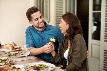 Present, happy couple and flower on dinner date for love, support and romance at valentines celebration. Giving, woman and man with gift at lunch for care, connection or laughing together in house