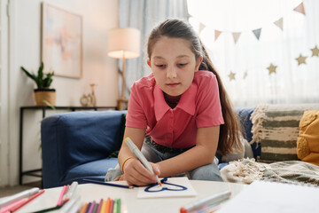 Young girl concentrating on drawing with crayons in warm, inviting living room setting. Focused expression showing her dedication to her artistic creation