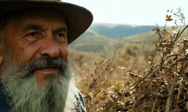 Wyschnięte, dzikie rośliny otaczają starszego mężczyznę o przenikliwym spojrzeniu, kt&oacute;ry nosi szeroki kapelusz i ma długą, siwą brodę, w scenerii leśnej.