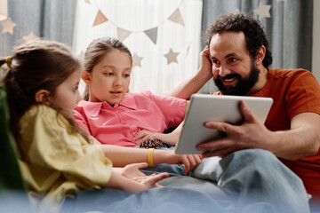 Father sharing enjoyable moment with daughters while using tablet on sofa, fostering bonding and interaction between them