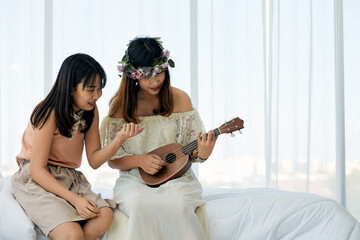Two women sitting together on bed, one playing ukulele while other offers guidance, with bright window in background