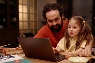 Father sitting with daughter while they engage in educational activity on laptop, sharing a moment of learning and connection at home