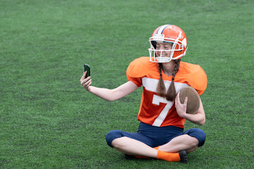 A girl or a young woman in a helmet and an American football uniform is sitting on the grass of the field with a ball, taking a selfie or video chatting.