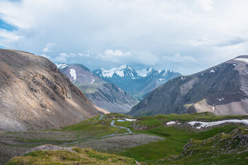 Scenic landscape with mountain creek and lake in green alpine valley against few big snowy pointy peaks far away under cloudy sky. Awesome aerial view to three large beautiful snow-capped peaked tops.