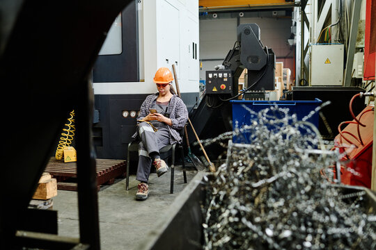 Worker in hard hat sitting on chair checking smartphone during break in factory setting surrounded by industrial equipment and scrap metal shavings