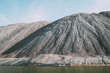 Rugged mountain slopes with textured surfaces under a bright blue sky, with a calm body of water at...