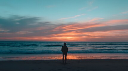 Fototapeta premium A solitary figure stands on the beach watching a serene sunset over the ocean, evoking feelings of tranquility and reflection. 