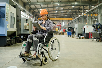 An industrial worker wearing a hard hat sitting in wheelchair conducting production check inside a...
