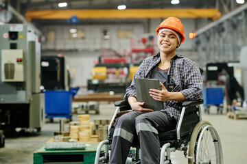 Woman in wheelchair wearing a hard hat and plaid shirt, smiling while holding clipboard in workshop, with industrial equipment and machinery in background - Powered by Adobe