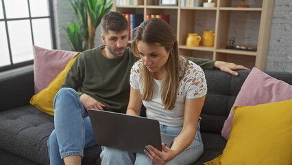 A woman and man sit closely on a couch in a cozy living room, attentively looking at a laptop...