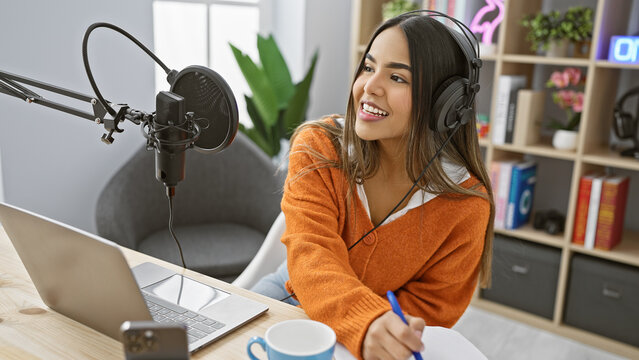A young hispanic woman records a podcast in a modern studio, equipped with a microphone, headphones, and a laptop.