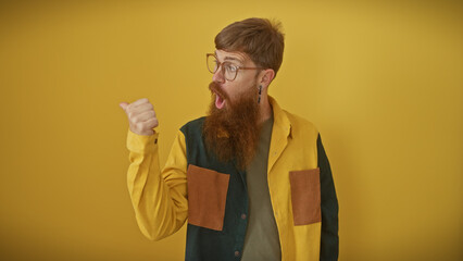 Surprised young adult redhead man with beard pointing thumb behind on isolated yellow background,...