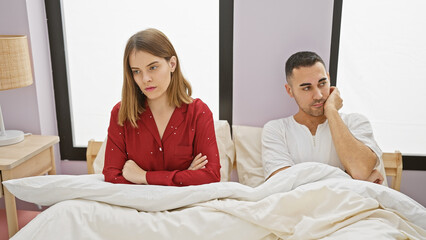 A woman and man sit apart in bed, portraying tension in a modern bedroom, hinting at relationship...