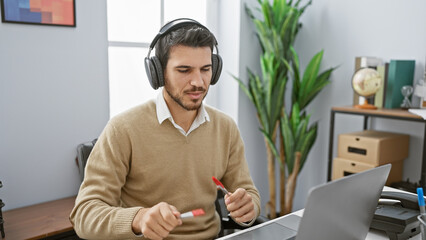 A focused hispanic man with headphones works at his tidy office desk, emanating professionalism and concentration.