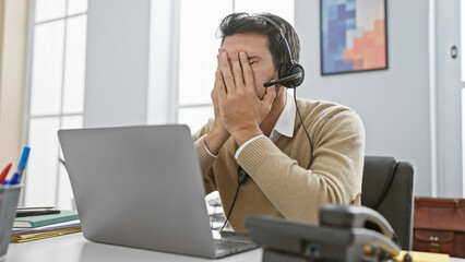 A stressed hispanic man with a beard wearing a headset covers his face at a modern office.