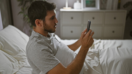 A handsome young hispanic man lying in bed while intently using a smartphone in a well-appointed bedroom