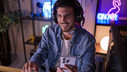 Handsome hispanic man with beard enjoying gaming in a dark room at night, wearing headphones and using a smartphone.