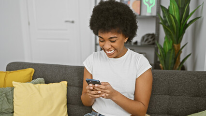 Smiling african american woman using smartphone in a cozy living room with modern decor.