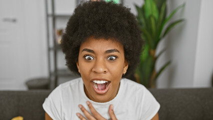 Excited african woman with curly hair in a white shirt surprised in a modern living room.