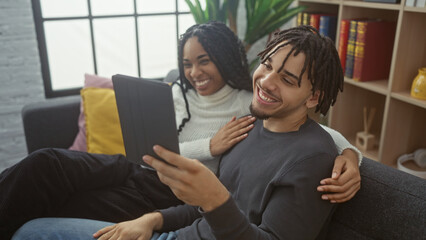 A joyous couple enjoys digital content on a tablet while cozily lounging in a modern apartment's living room.