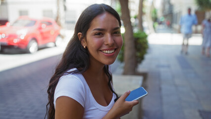 Young woman holding a phone and smiling on a city street, exuding happiness and vibrancy in an...