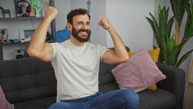A cheerful bearded man sits victoriously in a modern living room, exuding happiness and success.