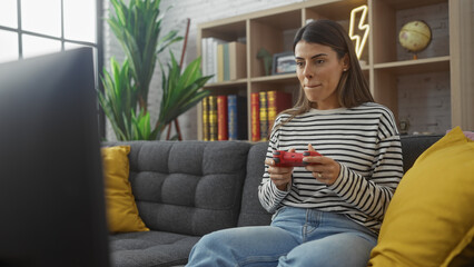 Obraz premium Hispanic woman gaming on a couch with controller in a living room with plant and bookshelf.