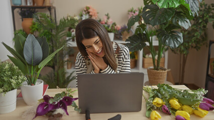 A cheerful young hispanic woman enjoys a video call at home surrounded by fresh flowers and indoor plants.