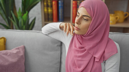 A thoughtful young woman wearing a pink hijab poses indoors, reflecting tranquility and elegance within a home setting.