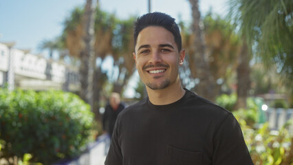 Handsome young man with a beard smiling outdoors in a sunny park with greenery and palm trees.