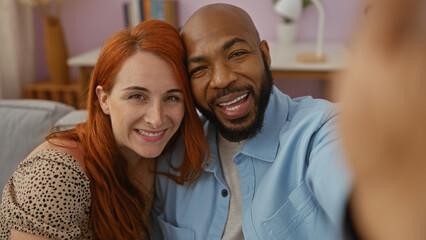 Interracial couple taking a selfie in their living room, capturing a moment of love and togetherness with a smiling man and woman inside their cozy home.