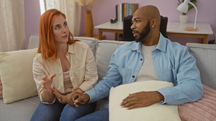 Interracial couple talking while sitting on a sofa in a cozy living room, demonstrating love and...