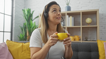 Pensive hispanic woman with headphones at home holding a yellow mug, resting on a grey couch against a white brick wall.