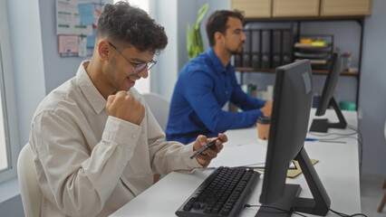 Men working in an office, one smiling at his phone while the other focuses on his computer,...
