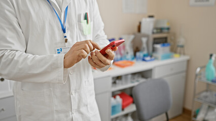 Hispanic man in lab coat texting on smartphone at medical facility.