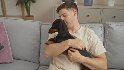 A young hispanic man holding a dachshund dog while sitting on a sofa in a cozy living room.