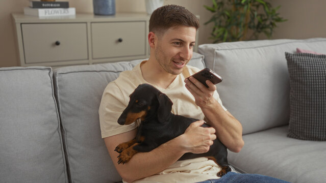 Young hispanic man in a living room holding a dachshund dog while sending a voice message on his smartphone - Powered by Adobe